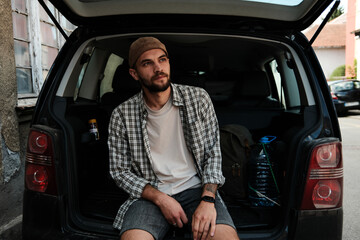 Young man sitting in open car trunk resting and thinking. Concept of freedom, travel pause, and urban lifestyle