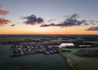 peaceful countryside village is nestled among fields and forests, with a reflective lake nearby, all bathed in the soft, fading light of a colorful sunset sky, sildemow