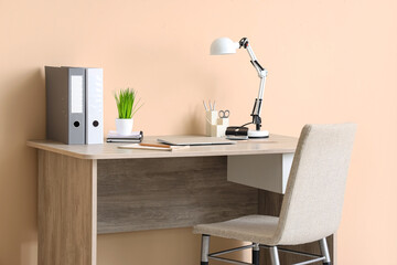 Modern workplace with desk lamp, houseplant, office supplies and chair near beige wall in room