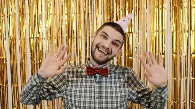Joyful man in checkered shirt and red bow tie smiling and waving at a birthday celebration against a shiny gold background. 4k horizontal footage - Powered by Adobe