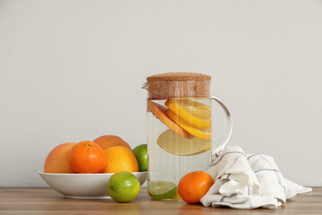 Jug with citrus lemonade and ingredients on table near white wall