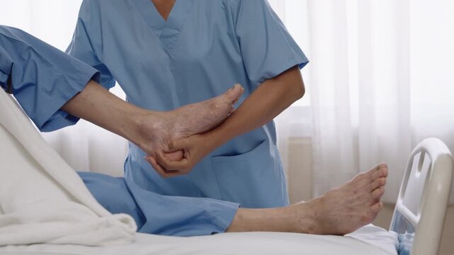 Asian nurse wearing blue scrubs helping elderly male patient with leg therapy on hospital bed, showing care and support during physical rehabilitation.