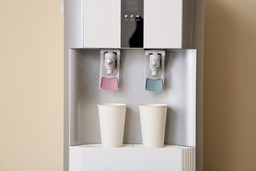 Modern water cooler with paper cups near beige wall in room, closeup