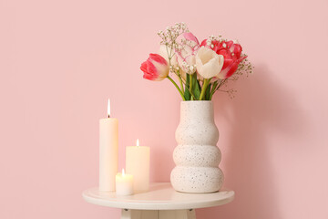 Burning candles and vase with flowers on table near pink wall in room, closeup