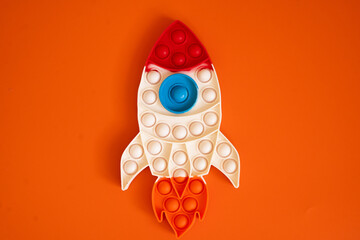 rocket shaped toy lies on a deep orange surface. Its design features red, blue, orange, and white silicone bubbles, making it a vibrant sensory and educational play tool for children.	