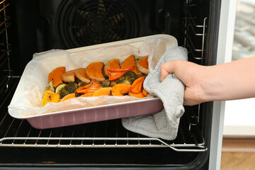 Woman taking baking tray with tasty baked pumpkin, eggplant, bell pepper and zucchini out of oven,...