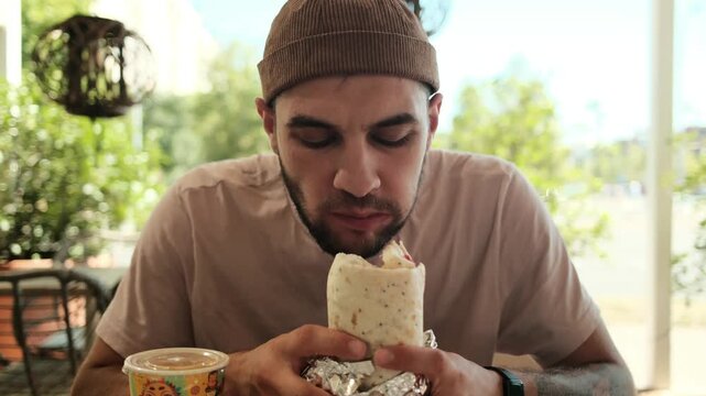 A young man enjoys eating a burrito while sitting at an outdoor Mexican cafe on a sunny day. Horizontal 4k footage