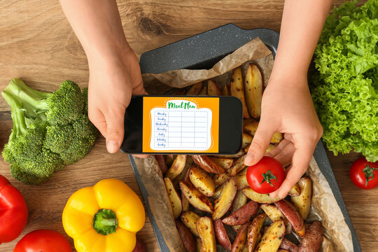 Young woman using meal plan app on mobile phone and holding tomato on wooden table in kitchen, closeup