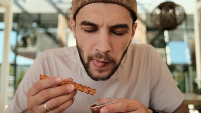 Man dipping fried churro stick into sauce and eating while sitting in Mexican cafe. Horizontal 4k footage