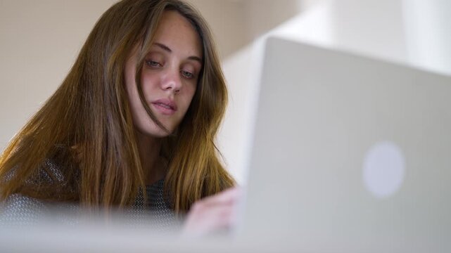 Woman using laptop for study and computer work at home indoor with long hair casual sweater student focused on screen learning homework research typing browsing web and smiling gently online