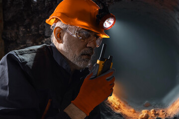 Man in a hard hat with a flashlight using a walkie-talkie in the darkness underground. Geology,...