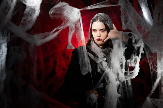 gothic woman with creepy vampire makeup trapped in spider web trying to scare say call me making phone gesture. Fantasy scary girl on red halloween background.