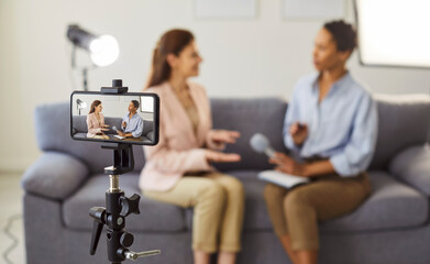 Smartphone camera on a tripod recording two women talking on a sofa during an interview, lighting,...