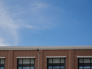 A cropped shot of the top of a modern industrial or commercial red brick building against a vast, bright blue sky with wispy clouds.