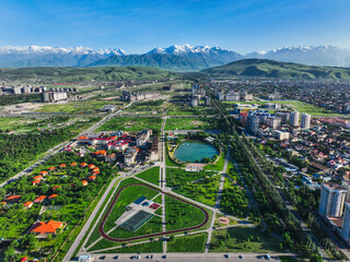 Wide aerial view of a large green park area with a running track and lake in Bishkek, backed by snow-capped Tian Shan mountains