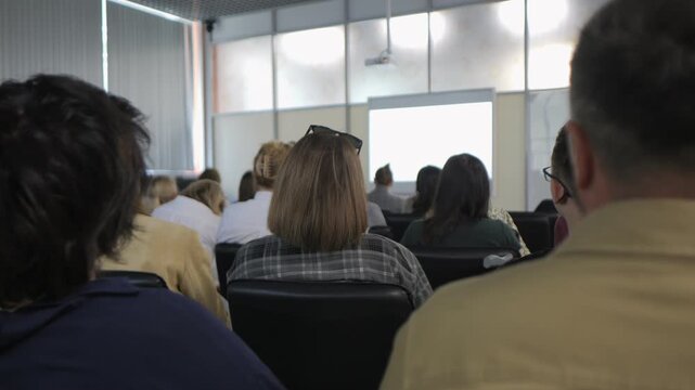 Group of people are sitting in a classroom. A man is sitting in the front row. A woman is sitting in the back row