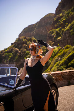 Elegant woman in black dress and headscarf posing by classic convertible car on a sunny coastal road, retro vintage style with luxury and glamour.