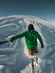 Point-of-view shot of a male skier in a green jacket carving through deep powder snow on a sunny mountain slope