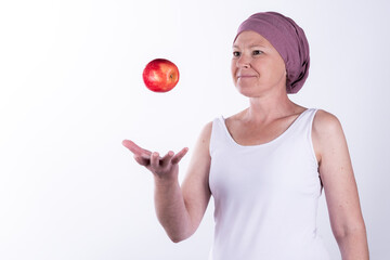 Smiling woman with a pink headscarf holding or tossing a red apple, symbolizing health, nutrition, and vitality during cancer recovery. Isolated on white background. Concept for advertising, press 
