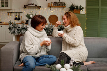 Two women enjoying warm beverages while sitting on a cozy sofa, wearing knitted sweaters, surrounded by festive decorations and a warm, inviting atmosphere for a joyful gathering