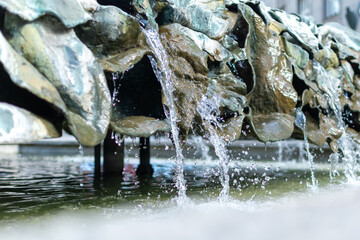 Close-up photo of a fountain where water cascades over a row of rough, organic-looking, and weathered metal or stone structures, splashing into the pool below