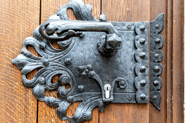 Close-up image of an elaborate, antique black wrought iron door handle and lock plate, mounted on a rustic wooden door with visible grain