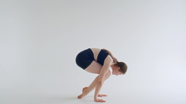 Young woman in sportswear practicing Bakasana pose in minimalist yoga studio, lifting feet off floor and balancing body weight on hands with knees supported on upper arms