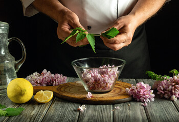 A chef carefully prepares a mix of fresh herbs and flower petals in a glass bowl. Slices of lemon and a carafe of water are also on the rustic wooden table, creating a serene cooking atmosphere