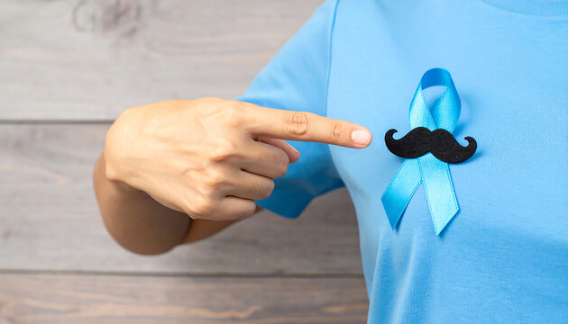 Close-up of a hand pointing to a symbolic light blue ribbon with a black mustache, representing prostate cancer and men's health awareness