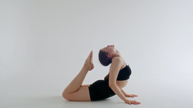 Flexible girl performing deep backbend pose with feet touching her head on floor against white background in minimalist yoga studio