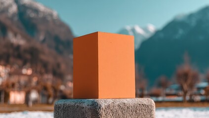 Orange geometric cube sits on stone pedestal with snowy mountains behind.