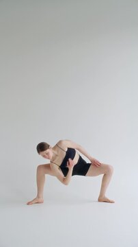 Girl performing advanced variation of compass pose, gripping lifted leg overhead and balancing on one arm in minimalist yoga studio with white cyc wall. Vertical clip
