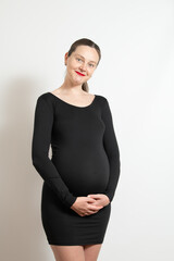 Obraz premium Expecting mother stands gracefully indoors wearing a black dress. She smiles warmly while gently cradling her baby bump, highlighting the beauty of pregnancy