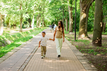 Fototapeta premium Front view of mother walking down the street with little son with sunglasses on sunny day in public green park.