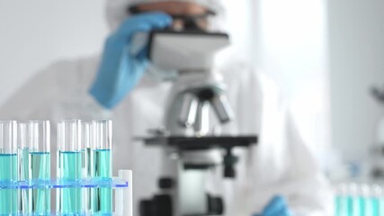 Medical scientist wearing protective suit using microscope while analyzing samples, conducting research in laboratory environment. Close up of test tubes with blue liquid - Powered by Adobe