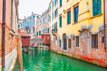 Narrow canal in Venice, Italy, with historic buildings on either side. A small arched bridge is visible in the distance, and the buildings are reflected in the green water