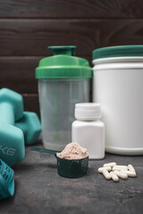 Scoop of whey protein powder with white amino acid capsules, fitness dumbbells, measuring tape, shaker bottle and protein container on dark surface, concept of sport nutrition and healthy lifestyle