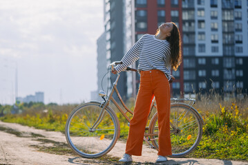 Young woman standing next to her city bicycle on a sunny day, looking up with closed eyes and a serene expression, embracing a moment of peace and freedom in an urban landscape