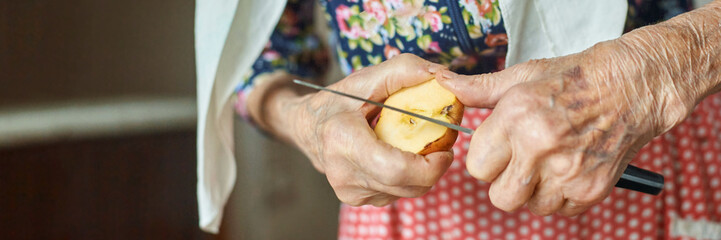 Elderly caucasian female peeling apple with knife in kitchen, floral blouse and red apron visible.
