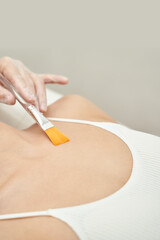 Hand applying beauty treatment brush on female chest in white top with soft light.