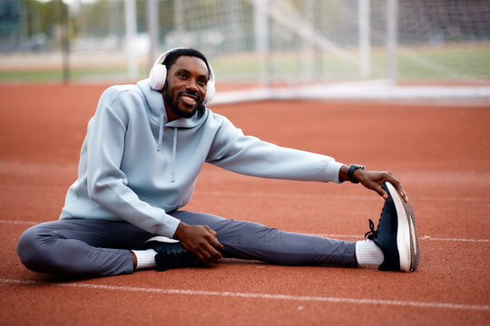 Happy black man stretching on running track with headphones