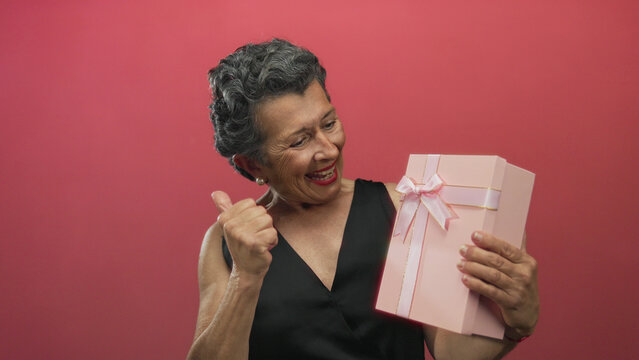 Senior woman with grey hair gives thumbs up while holding gift box against red background, showcasing joy and celebration.
