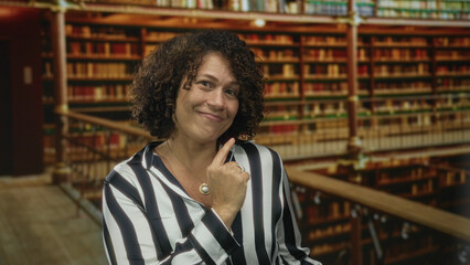 Woman smiling broadly pointing finger at bookshelf in building with curly hair; learning growth confidence.