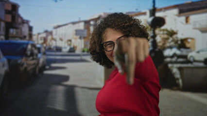 Woman with curly hair wearing red sweater and glasses poses peace sign by eyes on street while smiling under sunlight; playful joy.