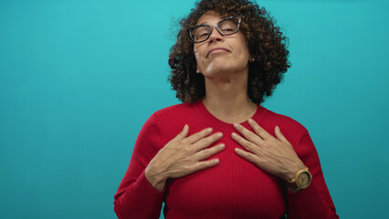 Woman adjusting her hands at neck in a turquoise studio wearing a red sweater and glasses;...