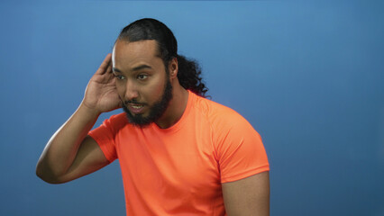 Man cupping hand to ear and leaning forward in studio against blue wall wearing orange shirt;...