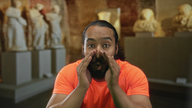 Man cupping hands around mouth shouting toward stone statues in museum building, orange shirt visible; surprise museum announcement.