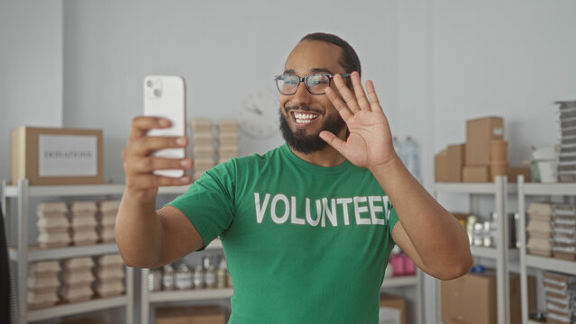 Man in green volunteer shirt smiling and holding a smartphone while giving thumbs up in a donations center building, surrounded by shelves and boxes; community support hope.