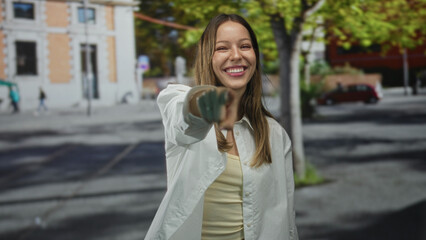 Fototapeta premium Young woman pointing finger at camera and smiling on street with building and trees in frame; playful invitation.