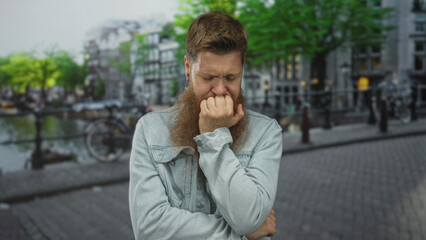 Man biting fingernails on street under green trees near canal while crossing arms in denim jacket;...